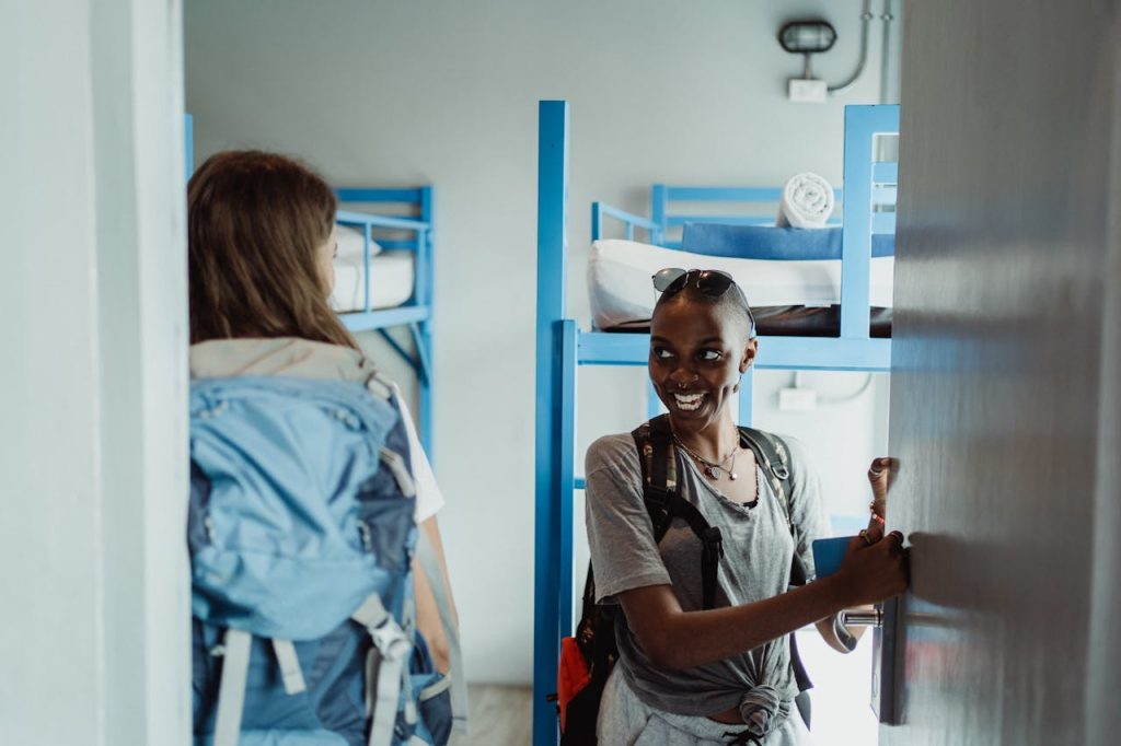 pexels photo 4907181 Two women backpackers entering a hostel dorm room with bunk beds, smiling and excited for their adventure.
