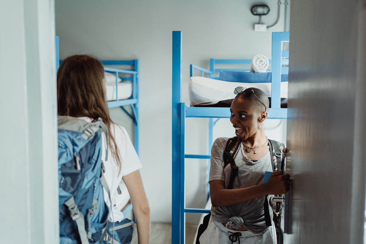 photography-service-03 Two women with backpacks walking into a bright hostel dormitory with bunk beds, ready for adventure.